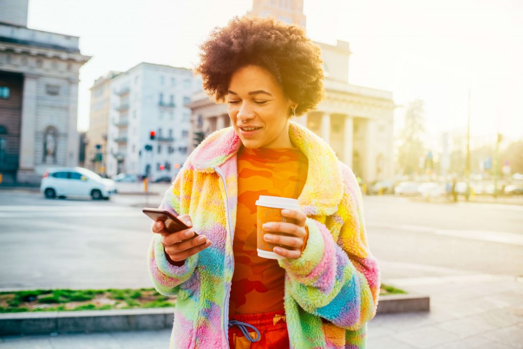 Woman walking through town looking at her phone while holding a disposable coffee cup (a common recycling contaminant)
