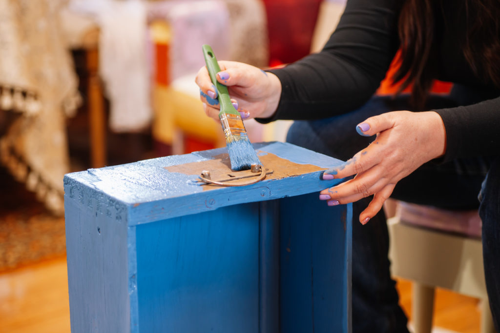 Closeup of woman with brush in paint-stained hands coloring wooden closet drawer in bright blue. Reuse of old things.